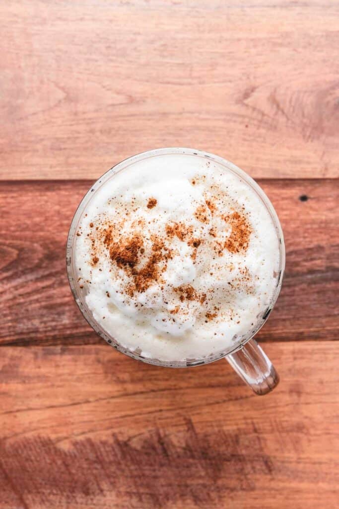 A glass mug of frothy Gingerbread Latte topped with cinnamon, on a wooden surface, viewed from above.