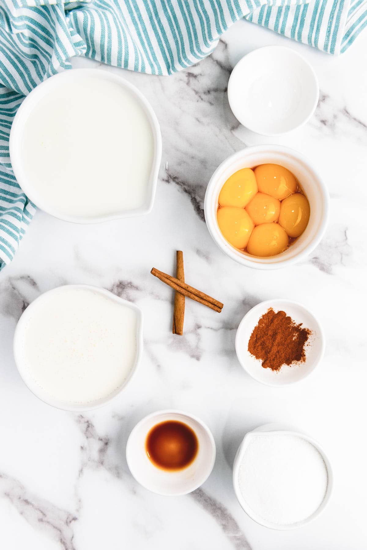 Egg yolks, milk, cream, cinnamon sticks, ground cinnamon, vanilla, and sugar on a marble surface with a striped towel&mdash;perfect ingredients for a creamy eggnog recipe.