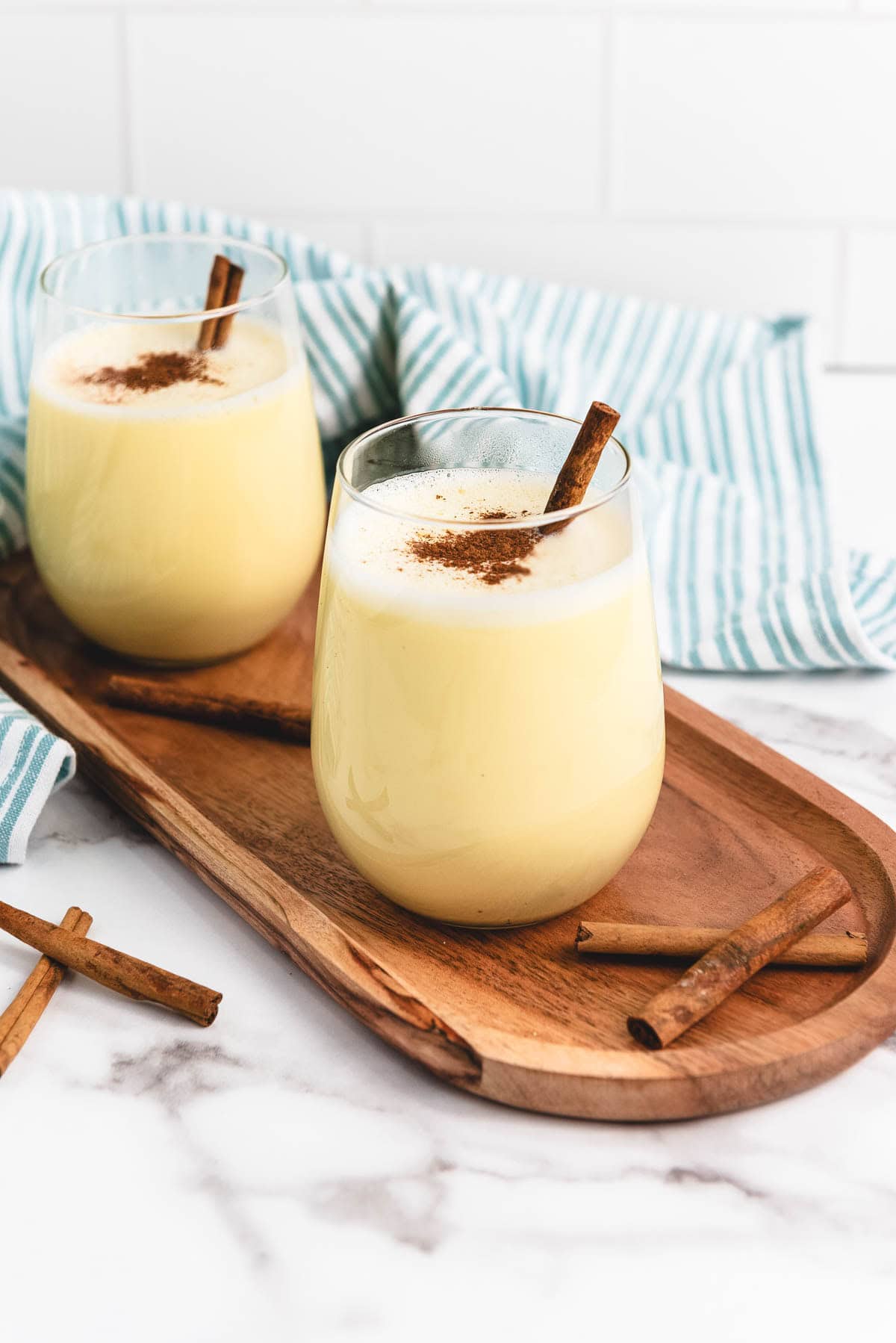 Two glasses of creamy eggnog topped with cinnamon sticks and powder rest on a wooden tray, offering a delicious taste of homemade eggnog.