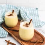 Two glasses of creamy eggnog with cinnamon sticks rest on a wooden tray, striped cloth in the background—perfect for enjoying your favorite homemade eggnog recipe.