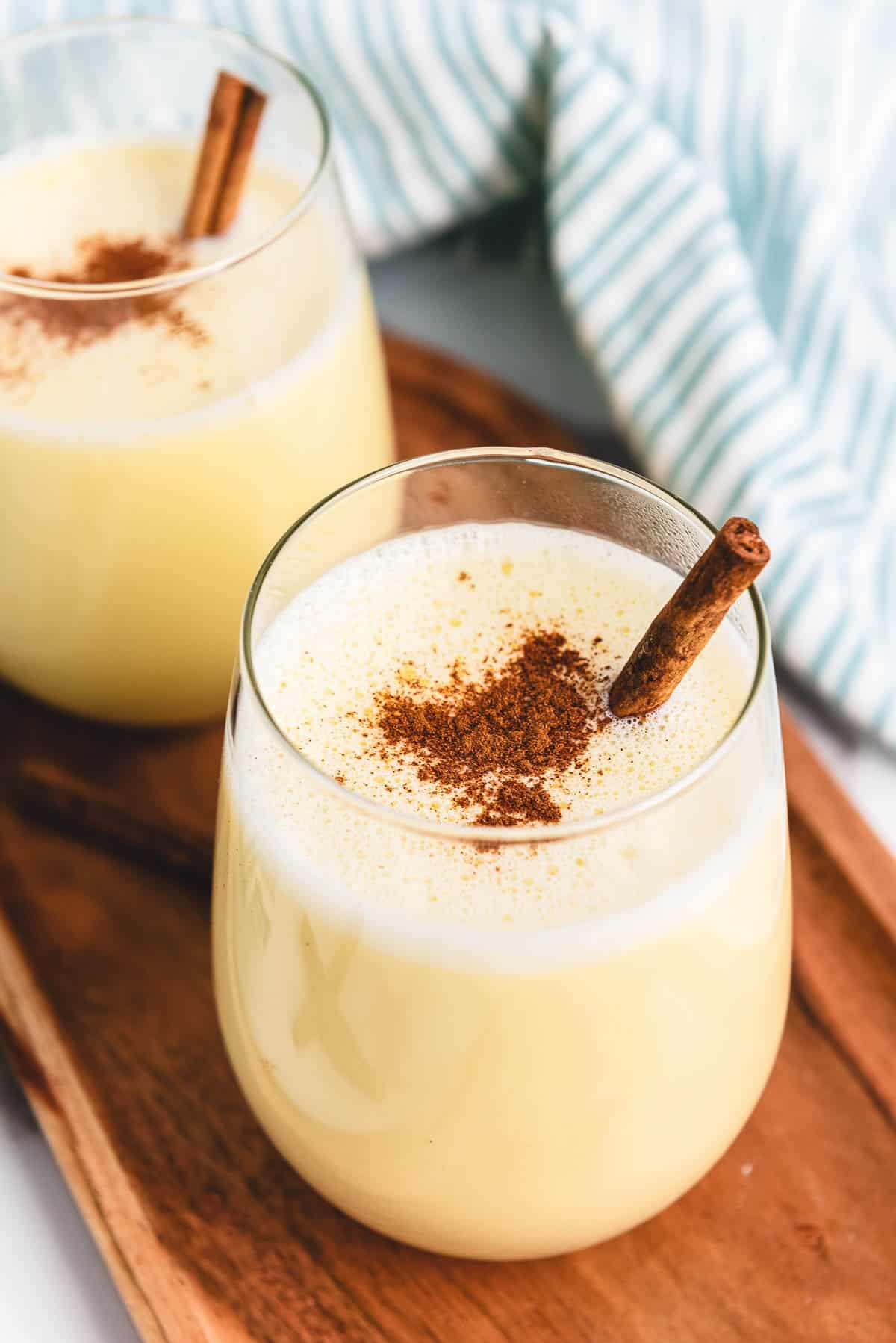 Two glasses of homemade eggnog topped with cinnamon and garnished with cinnamon sticks sit on a wooden tray, showcasing the perfect creamy eggnog recipe for the holidays.