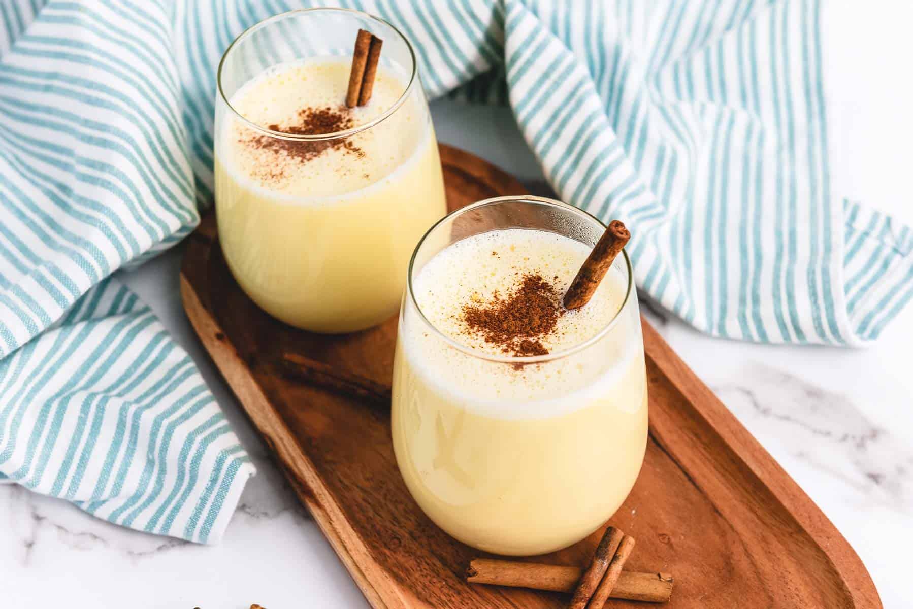 Two glasses of creamy eggnog with cinnamon sticks rest on a wooden tray beside a blue and white striped cloth, featuring a classic homemade eggnog recipe perfect for the holidays.