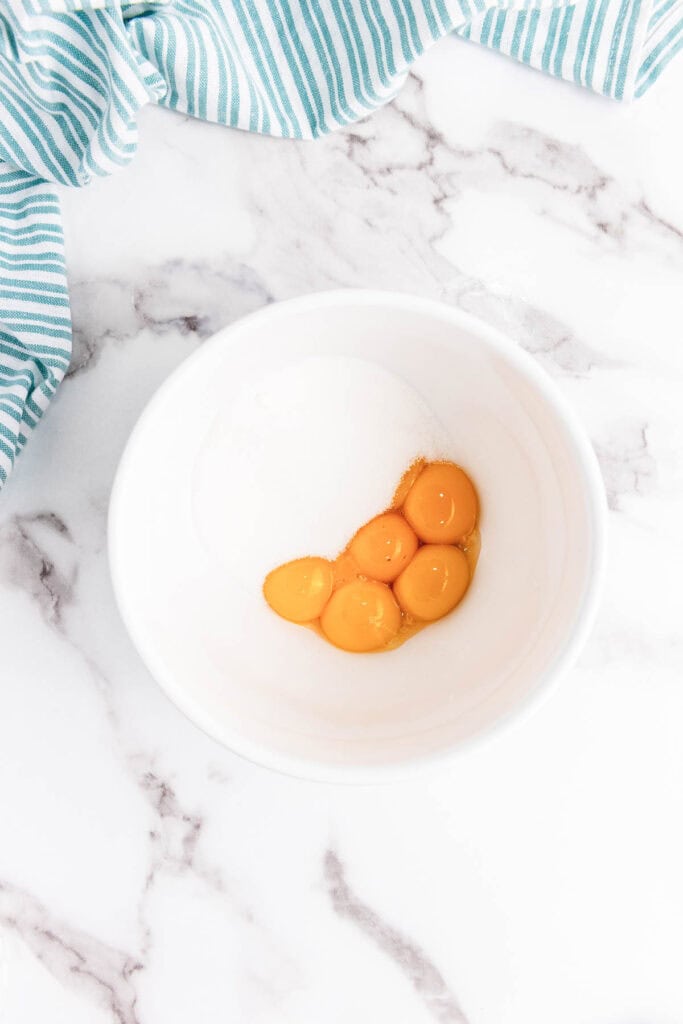 Five egg yolks and sugar in a white bowl on a marble countertop, ready to become creamy eggnog, next to a striped blue and white cloth.