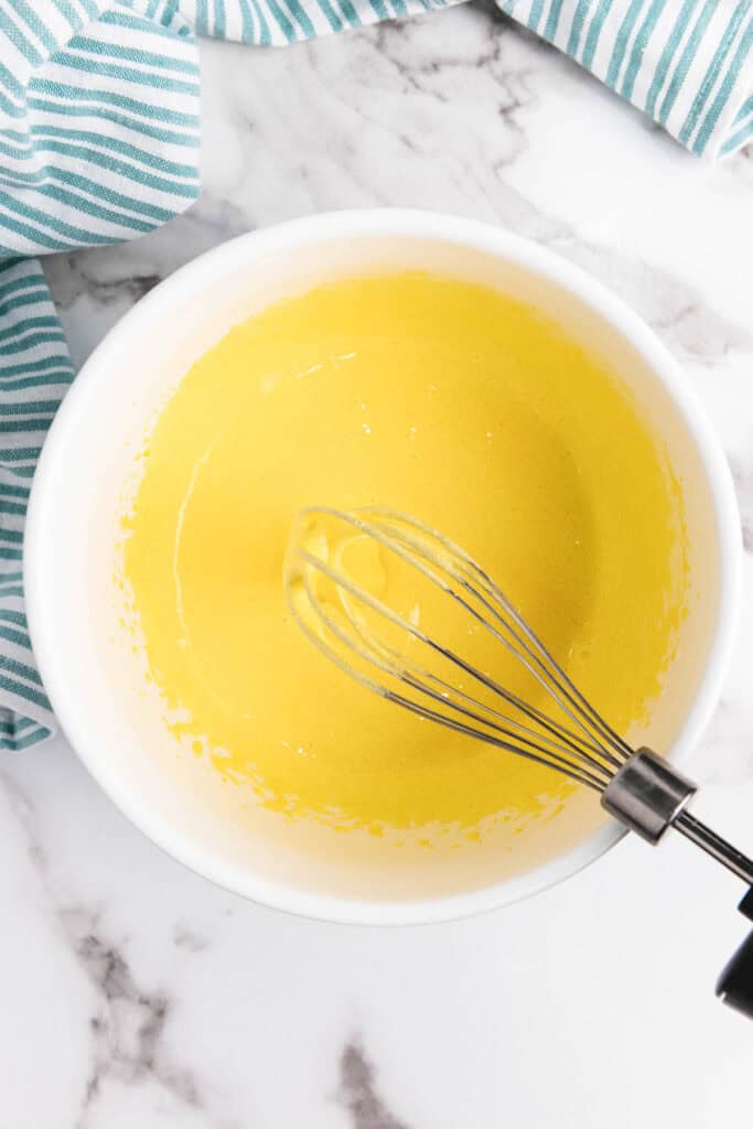 A metal whisk in a white bowl with yellow batter, perfect for preparing creamy eggnog, sits on a marble surface with striped towels nearby.