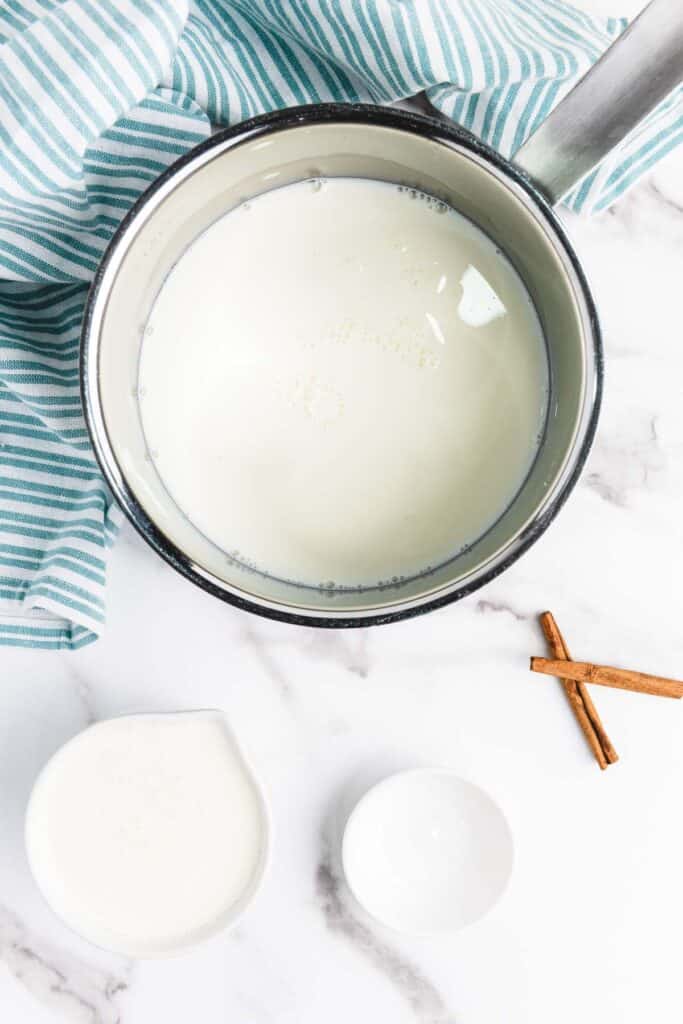A saucepan of milk with a striped towel, cream in a small pitcher, two cinnamon sticks, and a small empty bowl&mdash;perfect for starting your homemade eggnog or creamy eggnog recipe.