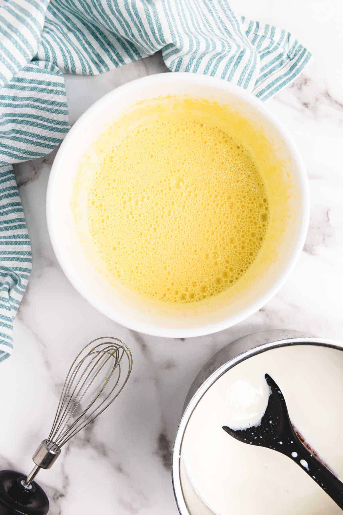 A bowl of beaten eggs next to a whisk, a bowl of cream, and a striped kitchen towel on a marble counter&mdash;perfect ingredients for a creamy eggnog recipe.