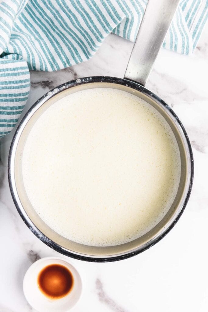 A saucepan of milk on a marble counter, with a small bowl of vanilla extract and a striped towel nearby&mdash;perfect for starting your homemade eggnog recipe.