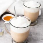 Two glass mugs of frothy London Fog tea lattes with spoons sit beside a dish of honey and a white cloth, inviting you to enjoy this Earl Grey-inspired treat.