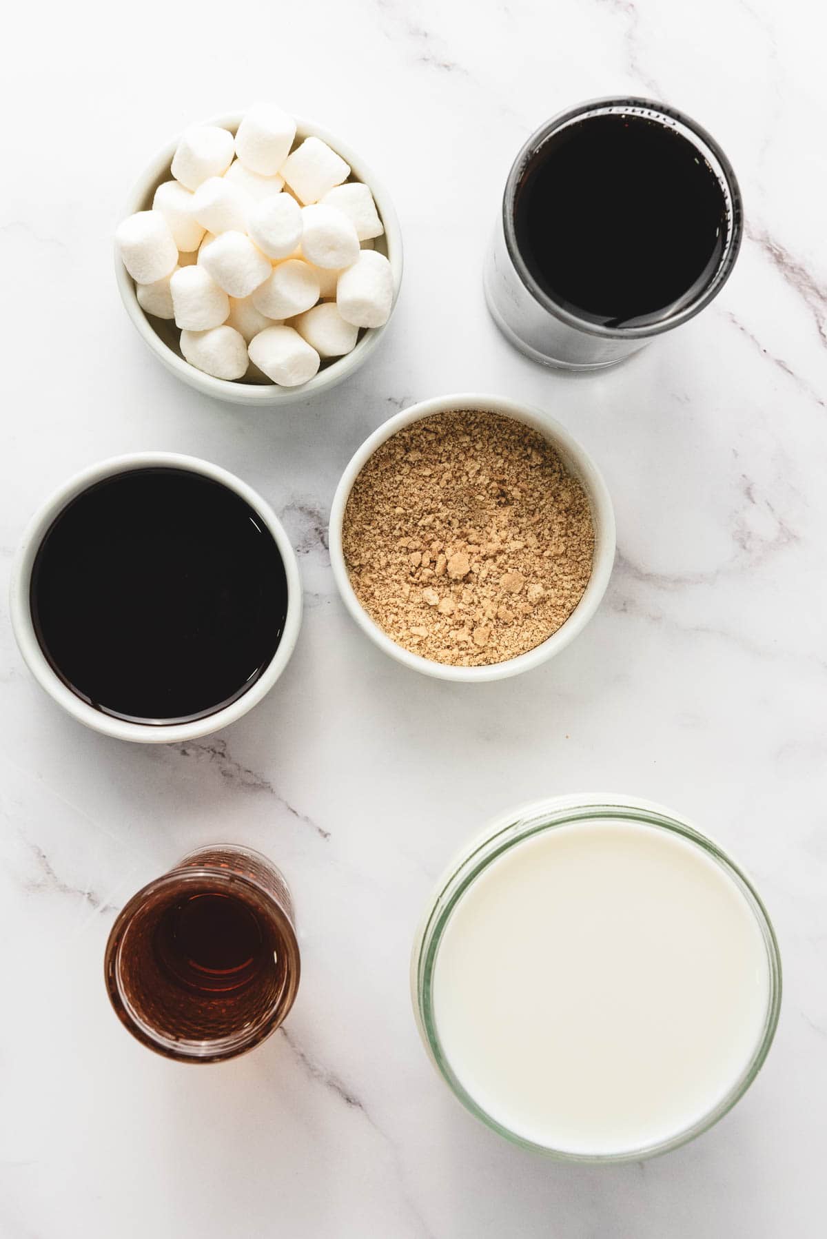 Bowls with marshmallows, chocolate syrup, crushed graham crackers, milk, vanilla, and coffee on a marble surface&mdash;perfect for crafting a delicious S&rsquo;mores Latte Recipe.