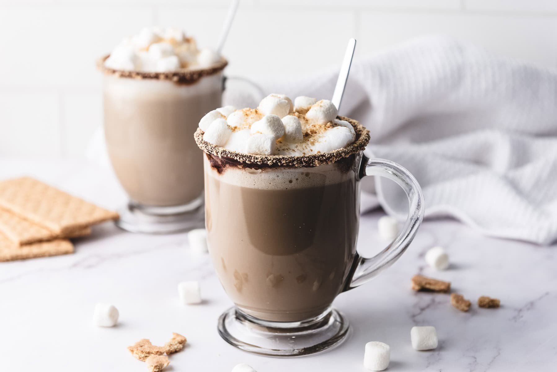 Two glass mugs of hot chocolate topped with marshmallows and graham cracker crumbs on a white surface&mdash;a cozy scene inspired by a classic S&rsquo;mores Recipe.