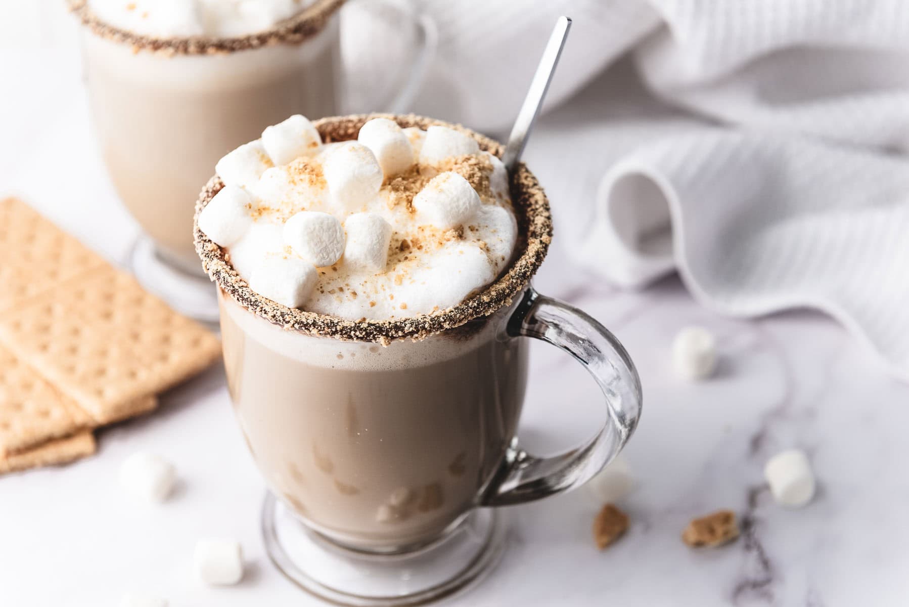 Glass mug of hot chocolate topped with marshmallows and graham crumbs, with graham crackers nearby&mdash;a cozy treat inspired by our S&rsquo;mores Latte Recipe.