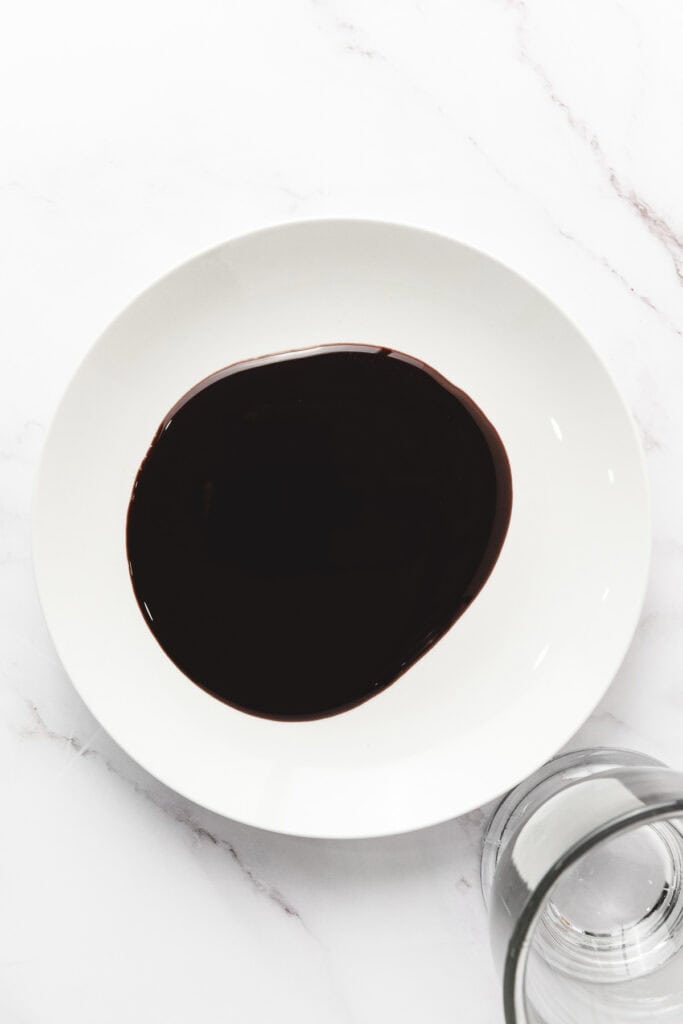 A white plate with a pool of dark chocolate sauce, perfect for a S&rsquo;mores Latte recipe, sits next to a clear drinking glass on a marble surface.