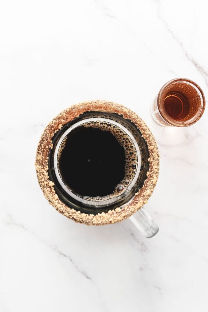 Top view of a mug with a crumb-coated rim filled with coffee, inspired by a S&rsquo;mores Latte Recipe, next to a small glass of brown liquid.