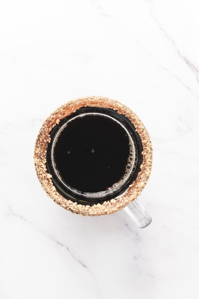 A glass mug of black coffee with a crumb-coated rim, viewed from above on a white marble surface&mdash;perfect inspiration for your next S&rsquo;mores Latte Recipe.