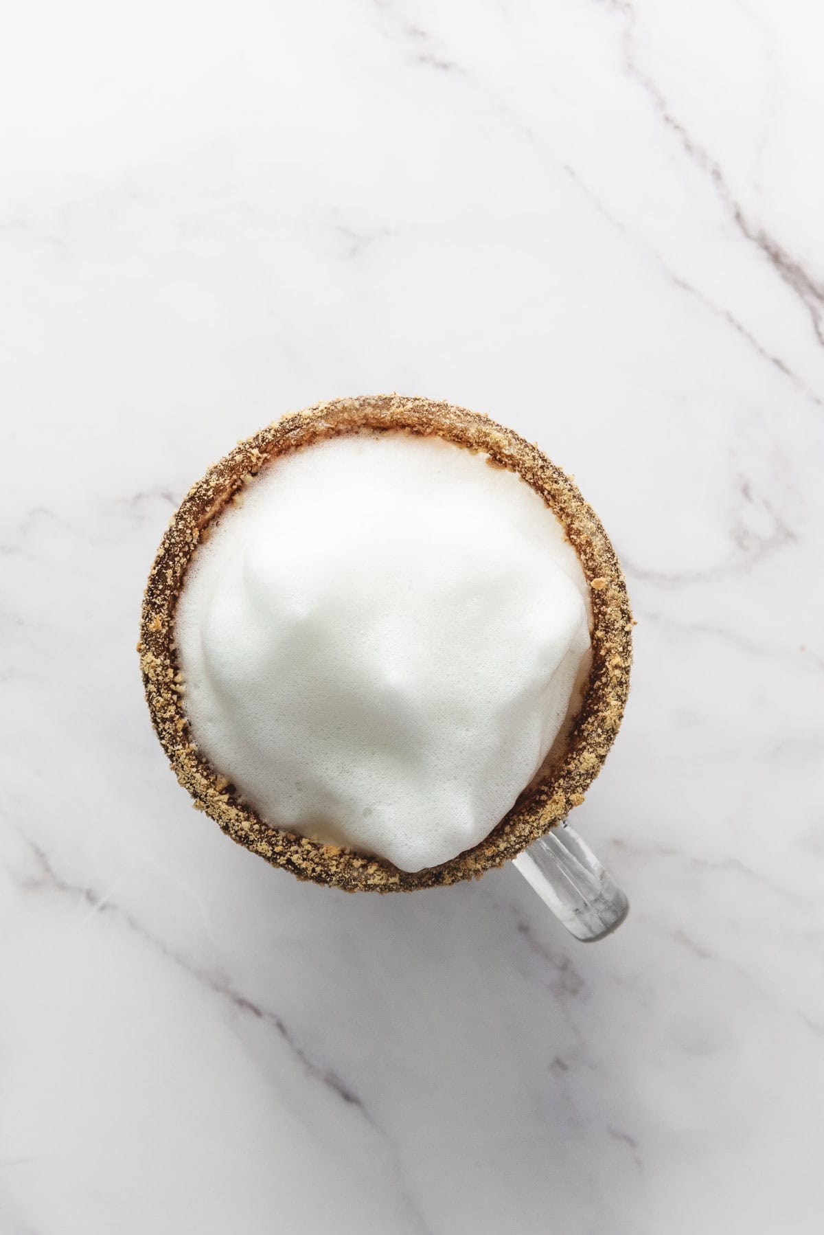 A glass mug of foamy S&rsquo;mores Latte with a crumb-coated rim sits on a white marble surface, viewed from above.
