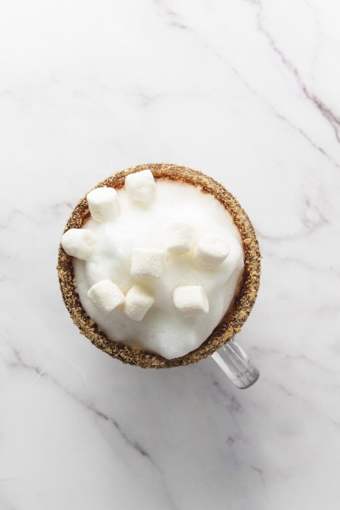 A mug of frothy S&rsquo;mores Latte topped with marshmallows and a graham cracker rim sits on a marble surface, perfect for anyone craving this cozy S&rsquo;mores Latte Recipe.