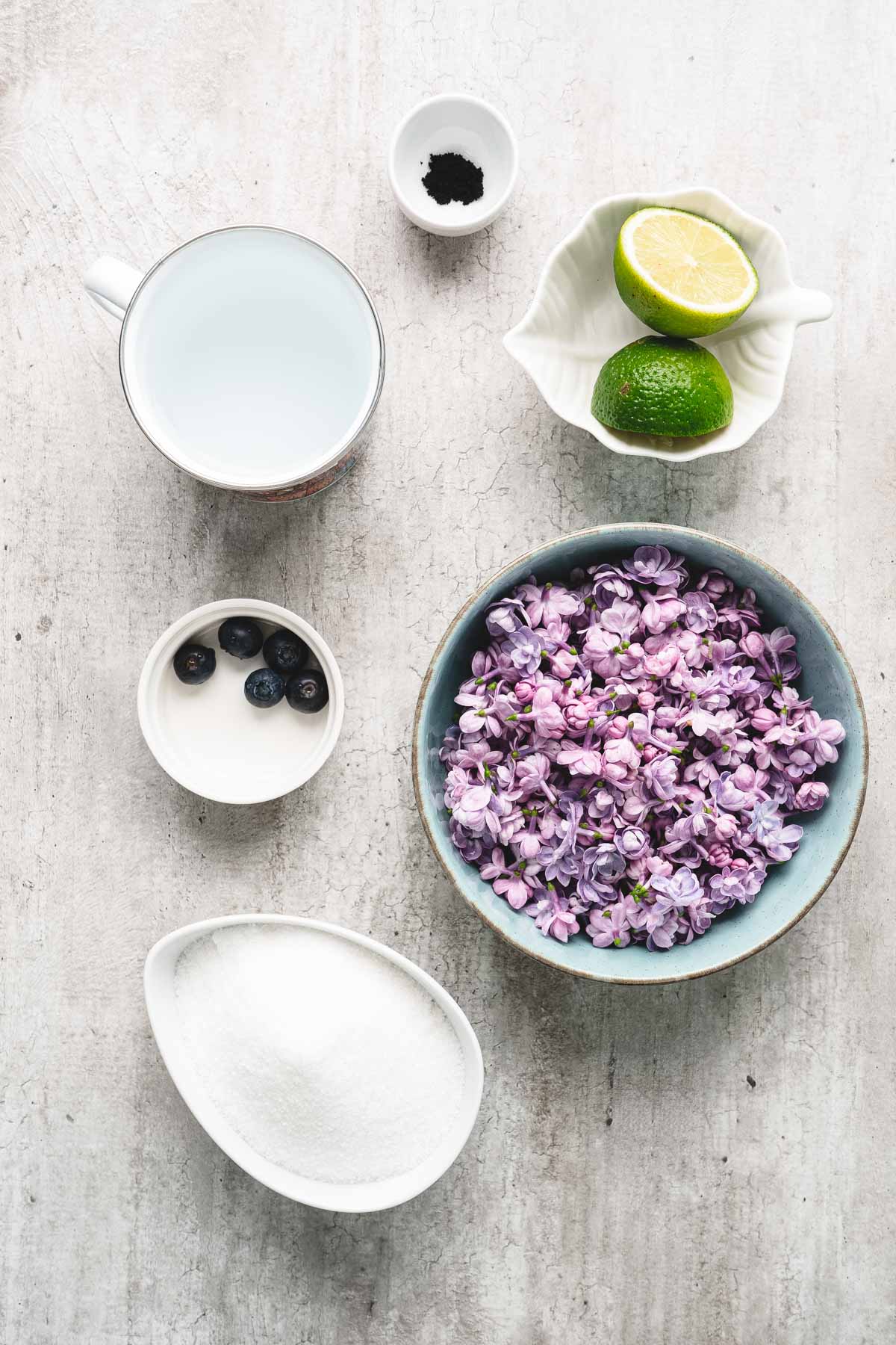 Bowl of lilac flowers, sugar, blueberries, lime, milk, vanilla, and a hint of lilac syrup on a light surface.