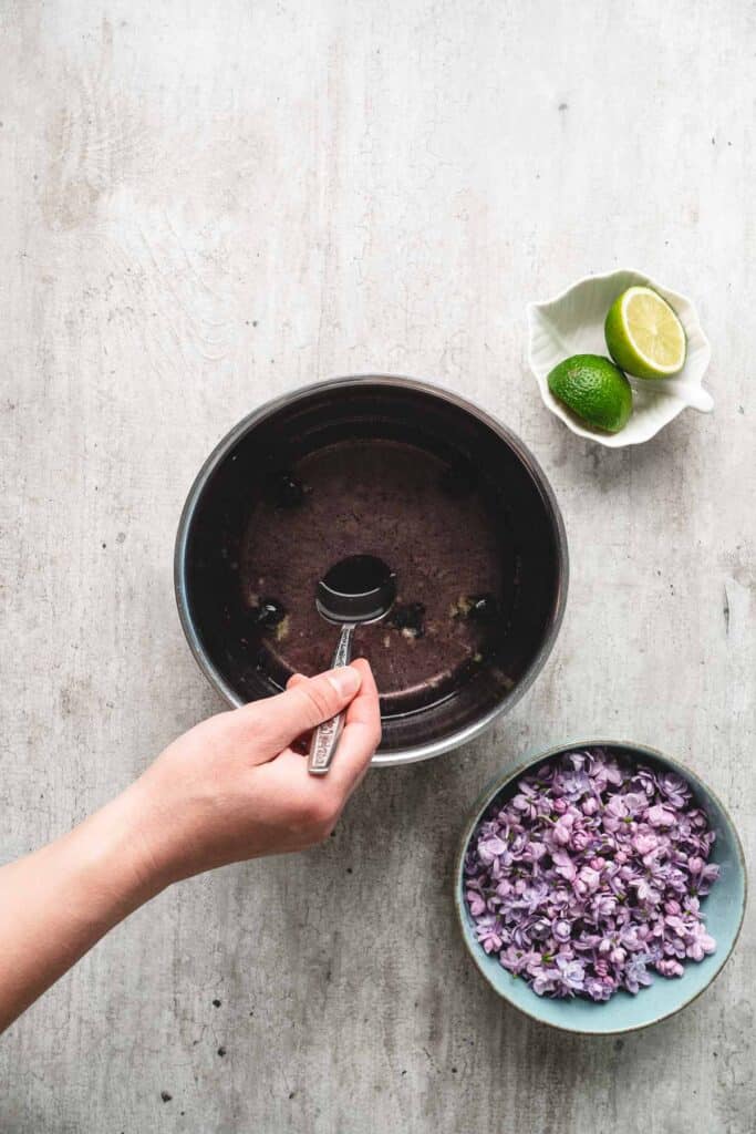 A hand holds a spoon over a bowl of dark liquid, with lilac syrup ready to pour. Nearby, a bowl of purple flowers and a halved lime add vibrant color to the scene.
