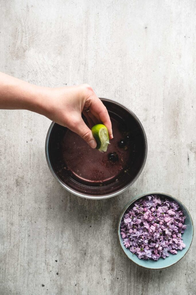 A hand squeezes a lime into a bowl of lilac syrup, next to a bowl of purple flowers on a light surface.