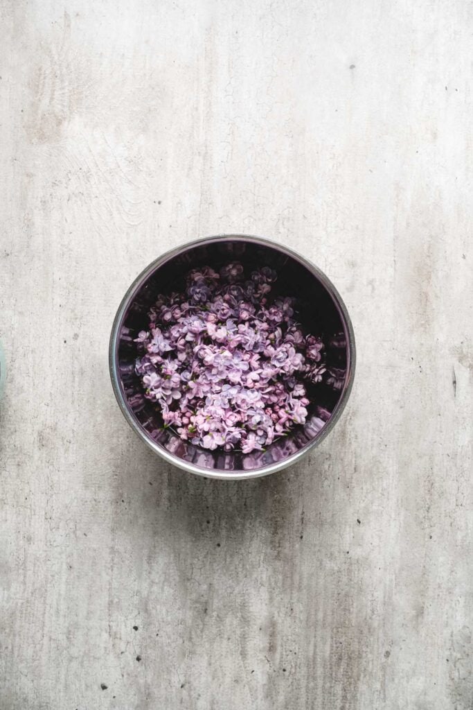 A metal bowl filled with small purple flowers sits on a light, textured surface, ready to be transformed into fragrant lilac syrup.