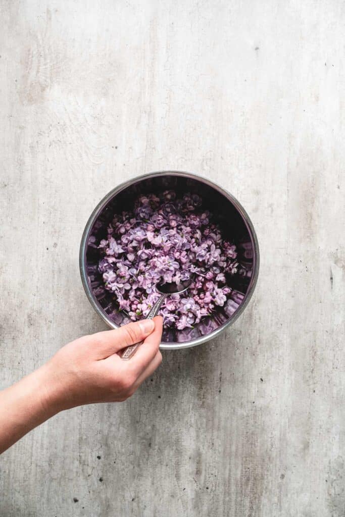 A hand stirs chopped purple flowers, soon to become lilac syrup, in a metal bowl on a light gray surface.