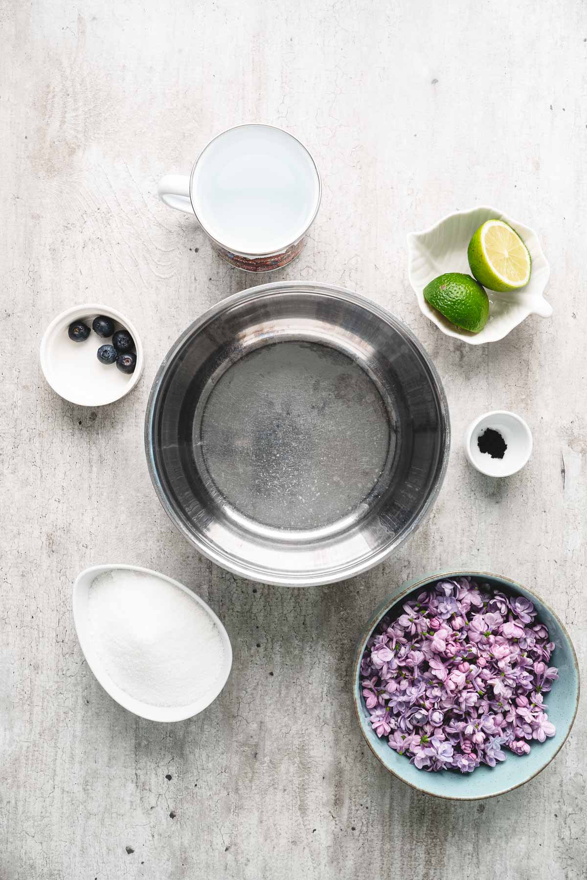 Metal bowl surrounded by lilac flowers, sugar, blueberries, lime, a cup of water, lilac syrup, and a small dish of black powder.