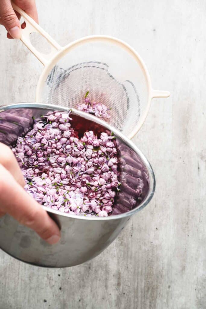 A hand pours purple lilac flowers from a metal bowl through a fine mesh strainer, preparing them for fragrant lilac syrup.