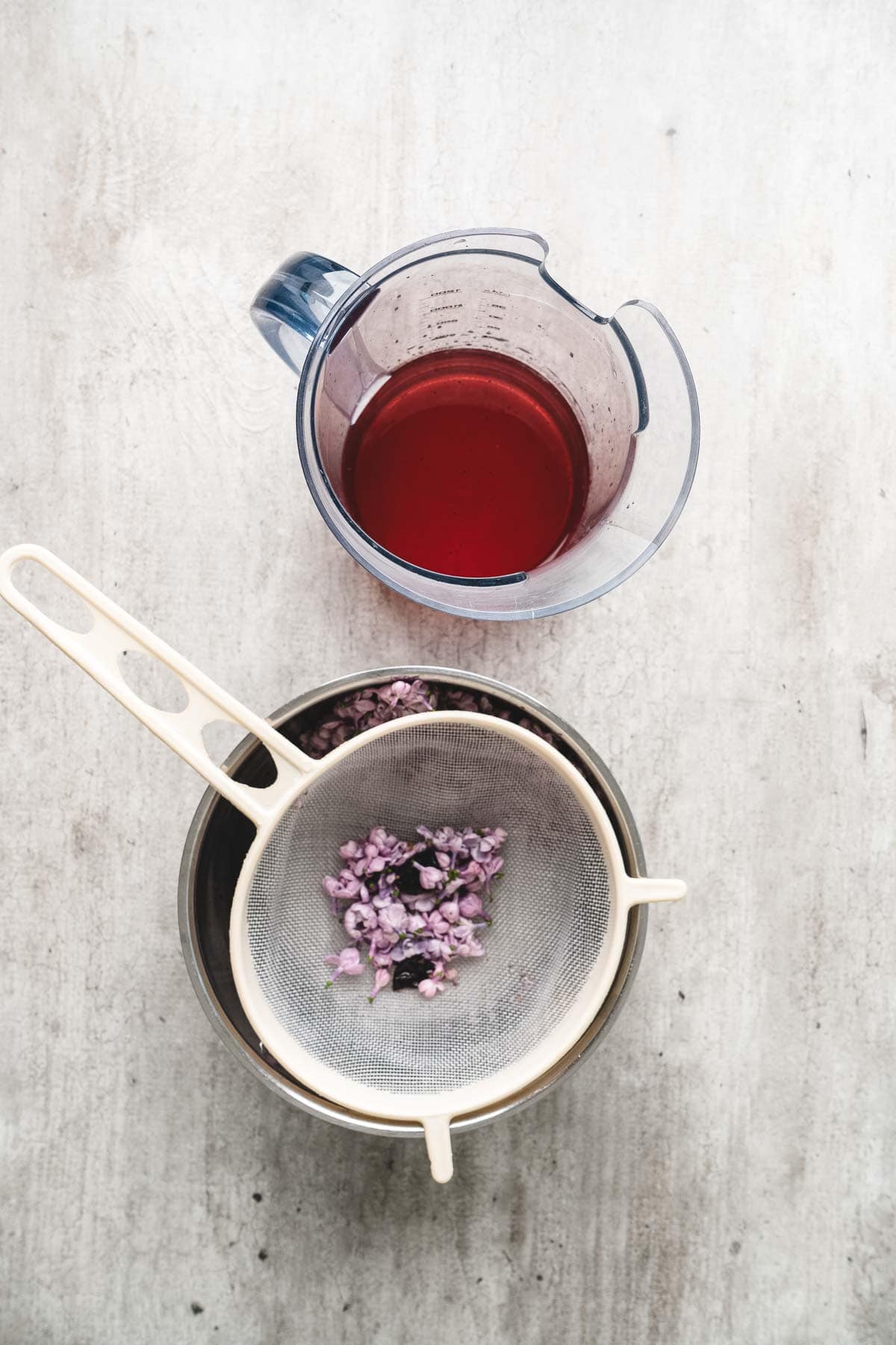A strainer with purple flowers, perfect for making lilac syrup, sits over a bowl beside a measuring jug filled with red liquid on a light surface.