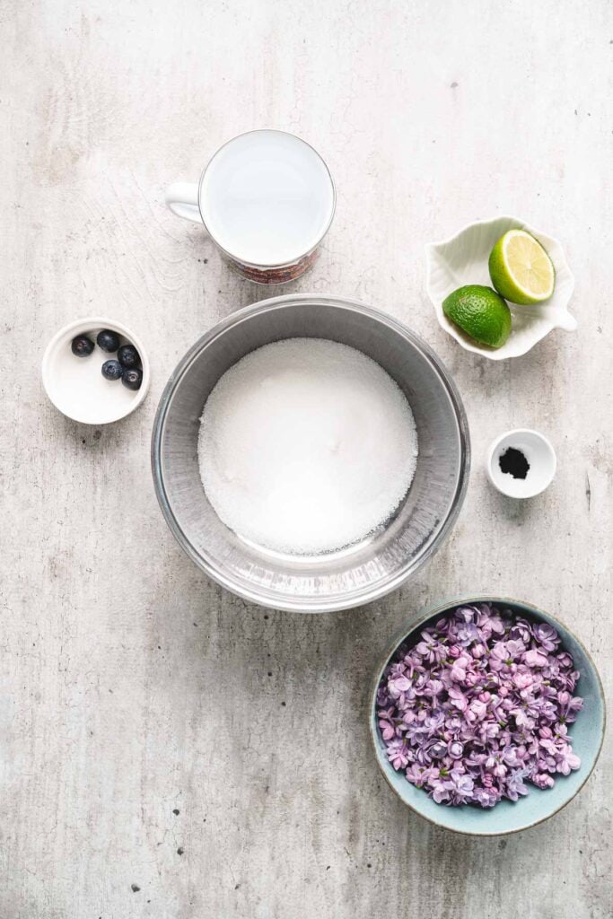Top view of sugar in a bowl, lilac syrup, lime halves, water, blueberries, and vanilla on a light surface.