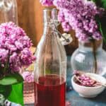 A glass bottle of red lilac syrup sits on a table, surrounded by vases of purple lilac flowers.