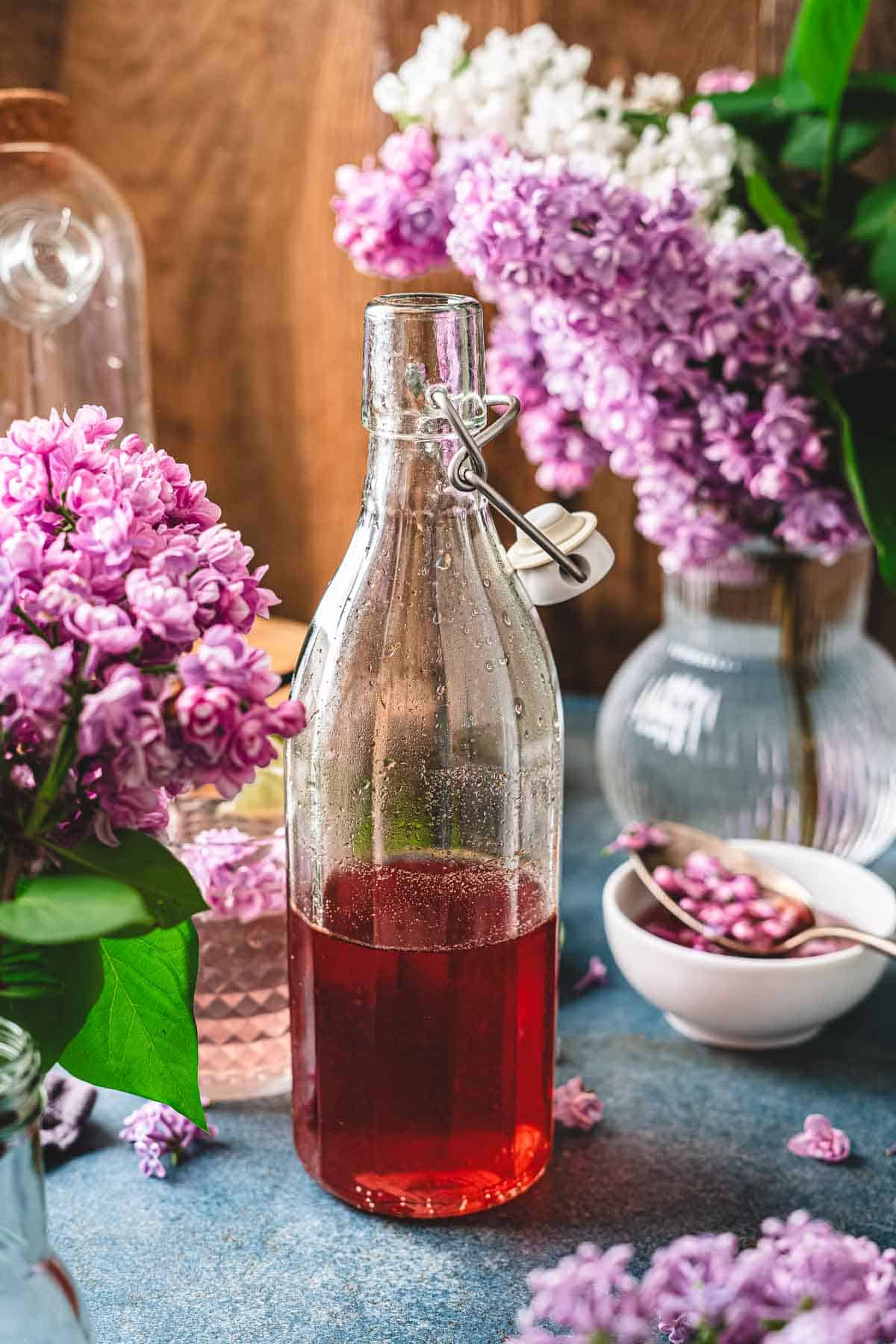 A glass bottle of red lilac syrup sits on a table, surrounded by vases of purple lilac flowers.