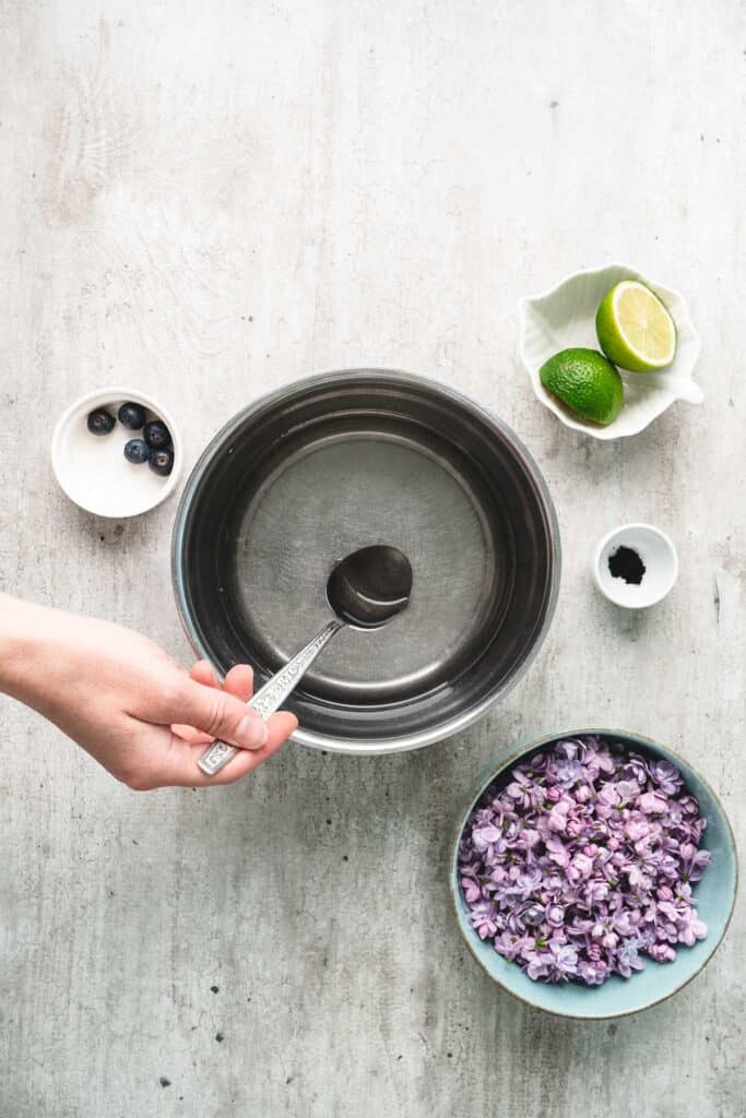 A hand holds a spoon over a pot; nearby are limes, blueberries, powder, and a bowl of purple flowers&mdash;perfect ingredients for crafting homemade lilac syrup.