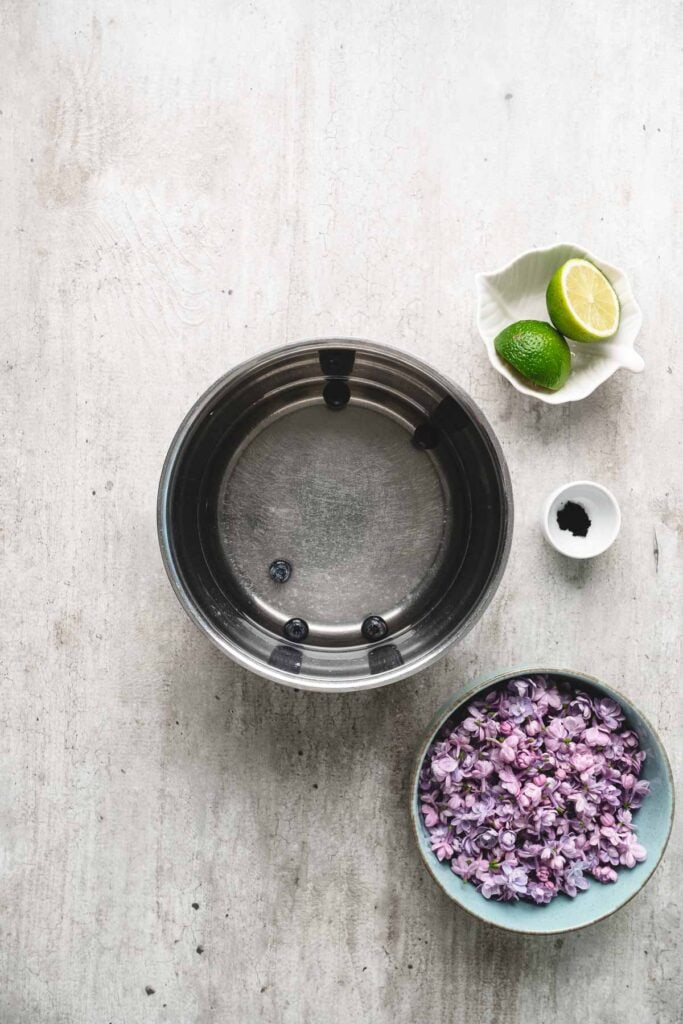 A metal bowl of water, a bowl of purple flowers ready for lilac syrup, a small dish of limes, and a tiny bowl with black powder.