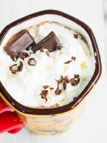 Mug of hot chocolate with whipped cream, chocolate pieces, and chocolate shavings, next to cinnamon sticks.