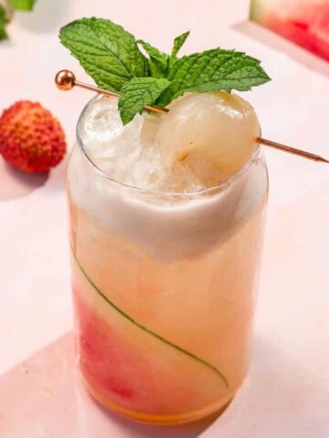 A tropical cocktail garnished with mint and lychee, surrounded by watermelon slices and lychees.