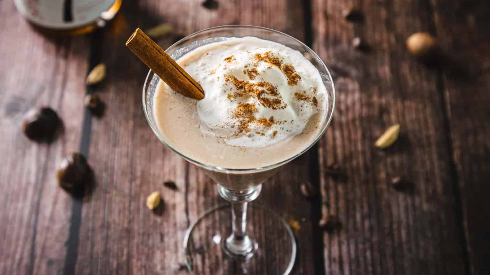 A creamy cocktail in a martini glass topped with whipped cream, cinnamon, and a cinnamon stick.