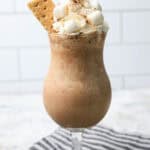 S'mores milkshake served in a stemmed glass, this iced mocha is topped with whipped cream, marshmallows, and a graham cracker on a striped cloth.