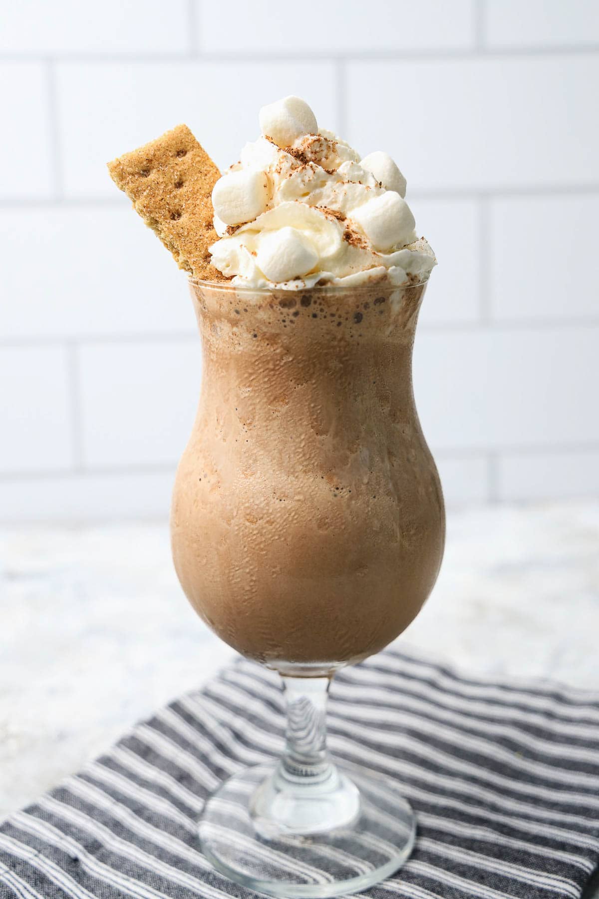 S'mores milkshake served in a stemmed glass, this iced mocha is topped with whipped cream, marshmallows, and a graham cracker on a striped cloth.