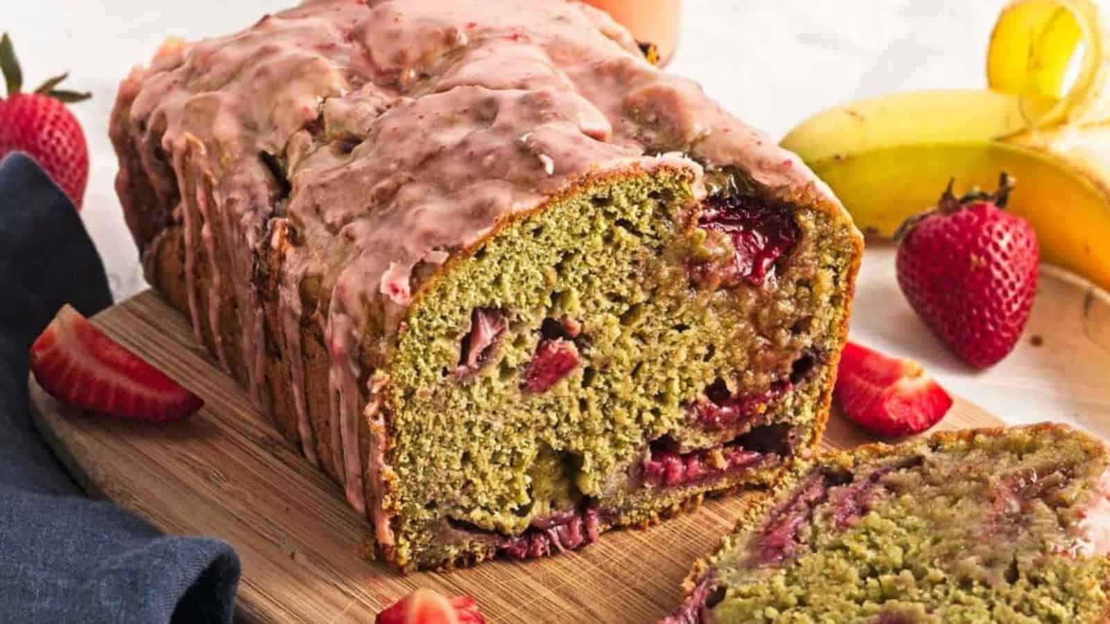 A sliced loaf of glazed strawberry banana bread on a wooden board, with fresh strawberries and bananas nearby.