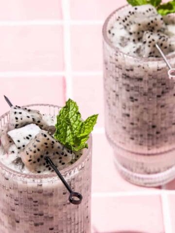 Two glasses of dragon fruit smoothie with mint leaves and straws on a pink tiled surface.
