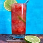 A tall glass of pink watermelon mocktail with lime slices and a straw, set against a bright blue background.