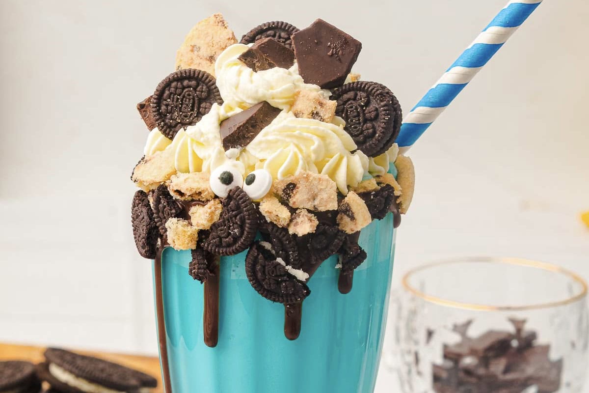 A blue glass of creamy Cookie Monster Milkshake is topped with whipped cream, cookies, chocolate, and a blue striped straw&mdash;the perfect milkshake recipe for any cookie lover.