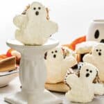 Ghost Muffins decorated with cream cheese icing shaped like ghosts, complete with chocolate eyes and mouths, are displayed on a white stand.