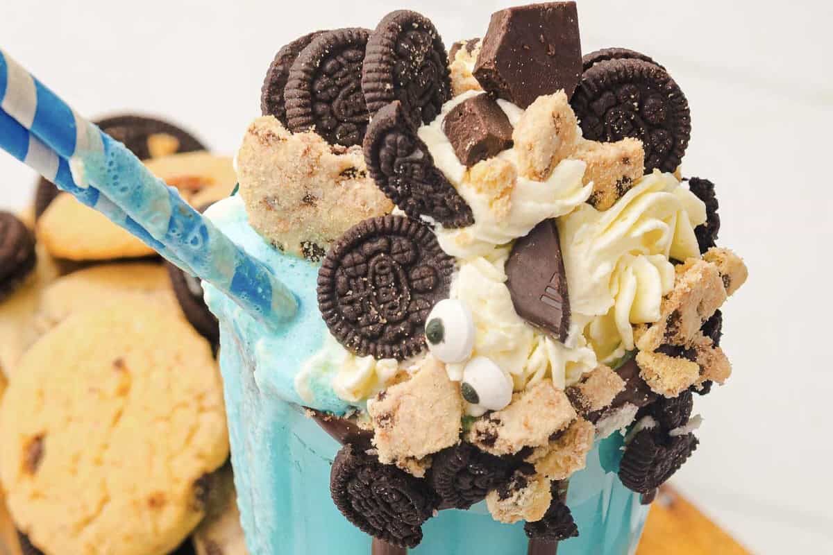 Blue cookie monster milkshake topped with whipped cream, mini cookies, chocolate pieces, and candy eyes, served with a blue straw.