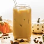 A glass of iced coffee with a straw, inspired by a Starbucks Copycat Oatmilk Shaken Espresso, surrounded by pumpkins and pecans on a wooden surface for a cozy Pumpkin Pecan vibe.