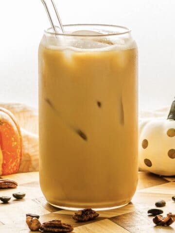 A glass of iced coffee with a straw, inspired by a Starbucks Copycat Oatmilk Shaken Espresso, surrounded by pumpkins and pecans on a wooden surface for a cozy Pumpkin Pecan vibe.