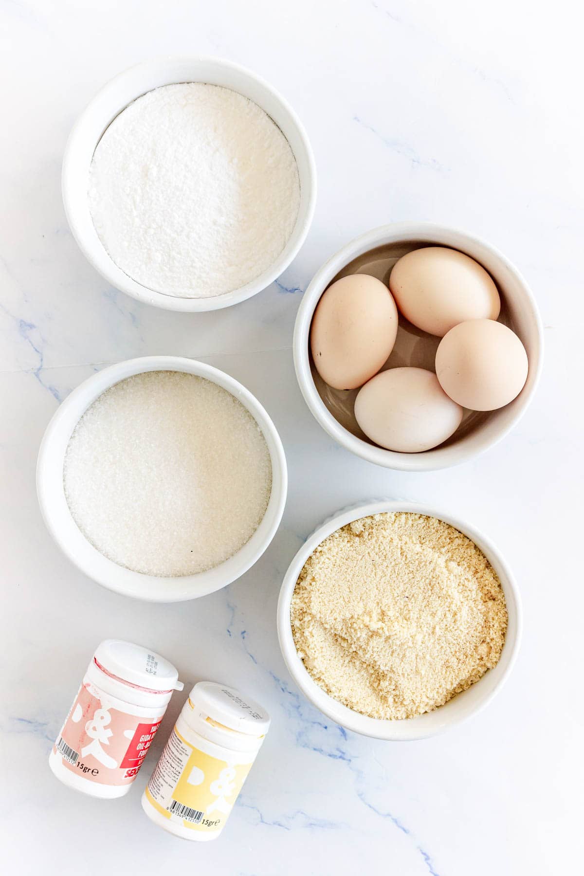 Five bowls with powdered sugar, whole eggs, granulated sugar, almond flour, and two containers of baking powder.