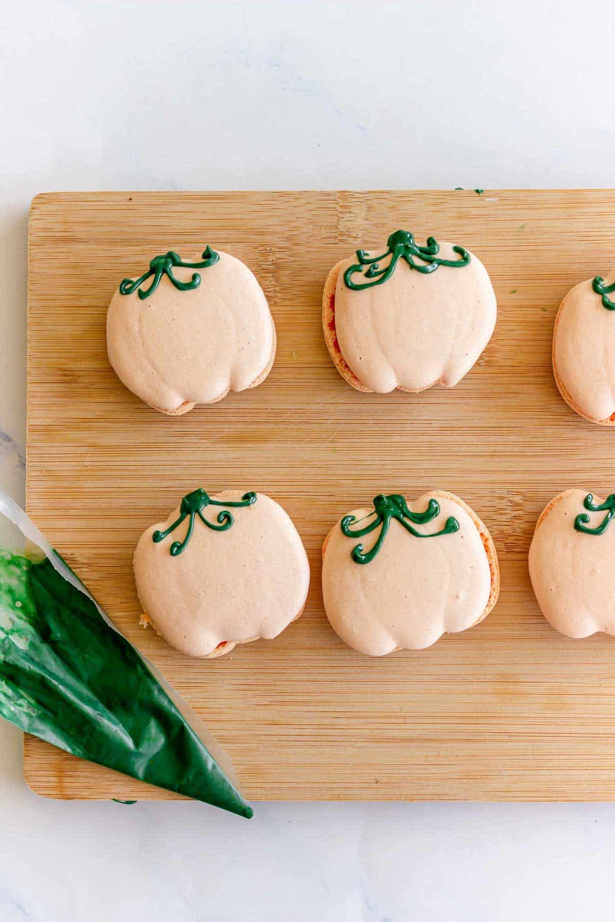 Pumpkin-shaped macarons with green icing on a wooden board, piping bag with green icing beside them.