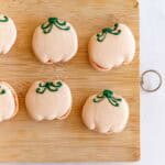 Six pumpkin-shaped macarons with green icing stems on a wooden board, next to a green icing bag.