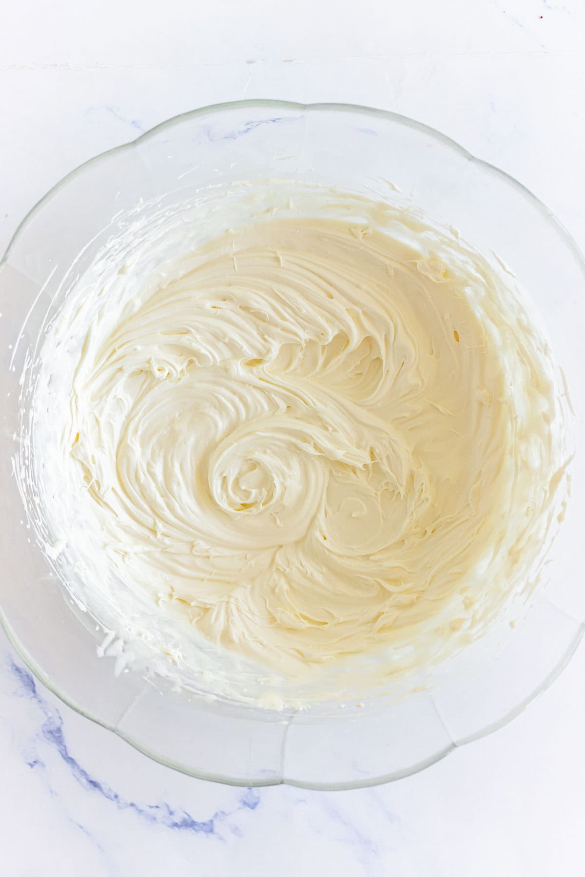 A glass bowl filled with smooth, creamy, light-colored frosting on a white surface.